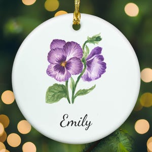 May include: A round white ceramic ornament featuring a watercolor illustration of purple pansy flowers with green leaves. The name Emily is printed in black cursive script below the floral design. A gold string is attached to the top hole.
