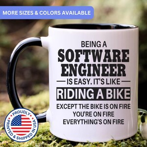 May include: A white ceramic mug with a black handle and interior, featuring black text that reads: BEING A SOFTWARE ENGINEER IS EASY. IT'S LIKE RIDING A BIKE EXCEPT THE BIKE IS ON FIRE YOU'RE ON FIRE EVERYTHING'S ON FIRE. Includes a circular flag emblem.