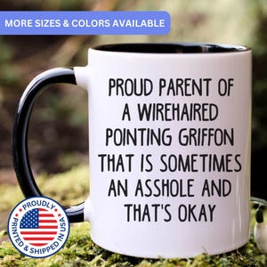 May include: A white ceramic mug with a black interior and handle. The mug features black text that reads: PROUD PARENT OF A WIREHAIRED POINTING GRIFFON THAT IS SOMETIMES AN ASSHOLE AND THAT'S OKAY. A circular blue, white, and red flag emblem is visible.