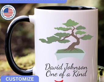 May include: A white ceramic mug with a black handle and interior rim. The front features a green bonsai tree graphic in a light blue pot. Below the tree, the text reads David Johnson One of a Kind in black script. A red arrow points to the text.