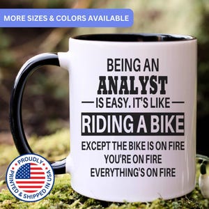 May include: A white ceramic mug with a black interior and handle. The mug features black text that reads: BEING AN ANALYST IS EASY. IT'S LIKE RIDING A BIKE EXCEPT THE BIKE IS ON FIRE YOU'RE ON FIRE EVERYTHING'S ON FIRE. A circular US flag emblem is included.