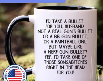 May include: A white ceramic mug with a black handle and interior rim. The mug features black text that reads: I'D TAKE A BULLET FOR YOU, HUSBAND. NOT A REAL GUN'S BULLET, OR A BB GUN BULLET, OR A PAINTBALL ONE. BUT MAYBE LIKE A NERF GUN BULLET? YEP, I'D TAKE ONE OF THOSE SONSABITCHES RIGHT IN THE HEAD FOR YOU!