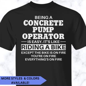 May include: A black short-sleeved t-shirt featuring white text that reads: BEING A CONCRETE PUMP OPERATOR IS EASY. IT'S LIKE RIDING A BIKE EXCEPT THE BIKE IS ON FIRE YOU'RE ON FIRE EVERYTHING'S ON FIRE. A blue banner at the bottom says MORE STYLES & COLORS AVAILABLE!