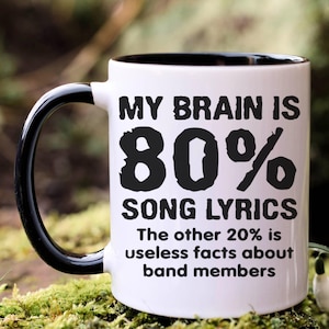 May include: White ceramic mug with a black handle and rim. The mug features the text "MY BRAIN IS 80% SONG LYRICS The other 20% is useless facts about band members" in black lettering. The mug is on a mossy surface.
