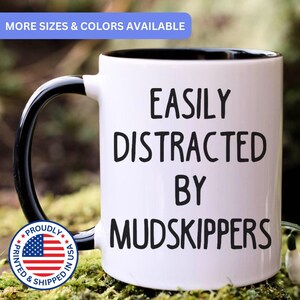 May include: A white ceramic mug with a black interior and a black handle. The front of the mug features the black text "EASILY DISTRACTED BY MUDSKIPPERS" in a sans-serif font. The mug has a smooth finish and a cylindrical shape.