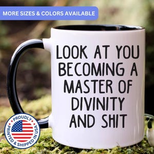 May include: A white ceramic mug with a black interior and a black handle. The exterior features black text that reads: LOOK AT YOU BECOMING A MASTER OF DIVINITY AND SHIT. The mug has a cylindrical shape and a smooth, glossy finish on the outer surface.