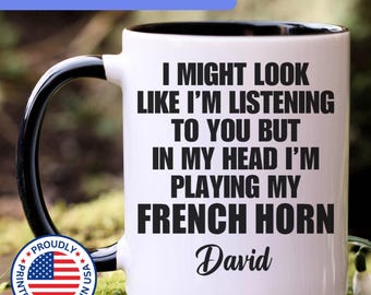 May include: A white ceramic mug with a black interior and handle, featuring the black printed text: I MIGHT LOOK LIKE I'M LISTENING TO YOU BUT IN MY HEAD I'M PLAYING MY FRENCH HORN, followed by the name David in a cursive script font.