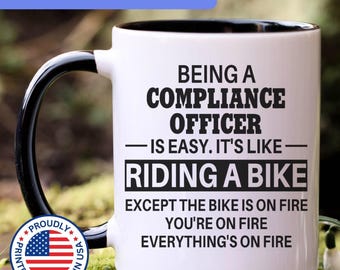 May include: A white ceramic mug with a black handle and interior rim. The mug features black text that reads: BEING A COMPLIANCE OFFICER IS EASY. IT'S LIKE RIDING A BIKE EXCEPT THE BIKE IS ON FIRE YOU'RE ON FIRE EVERYTHING'S ON FIRE.
