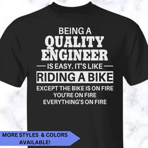 May include: A black short-sleeved t-shirt featuring white text that reads: BEING A QUALITY ENGINEER IS EASY. IT'S LIKE RIDING A BIKE EXCEPT THE BIKE IS ON FIRE YOU'RE ON FIRE EVERYTHING'S ON FIRE. The shirt has a crew neckline and a standard fit.