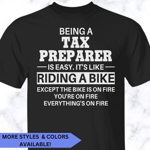May include: A black short-sleeved t-shirt featuring white text that reads: BEING A TAX PREPARER IS EASY. IT'S LIKE RIDING A BIKE EXCEPT THE BIKE IS ON FIRE YOU'RE ON FIRE EVERYTHING'S ON FIRE. The shirt has a crew neckline and a standard fit.