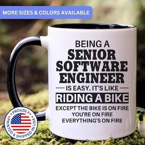 May include: A white ceramic mug with a black handle and interior, featuring black text that reads: BEING A SENIOR SOFTWARE ENGINEER IS EASY. IT'S LIKE RIDING A BIKE EXCEPT THE BIKE IS ON FIRE YOU'RE ON FIRE EVERYTHING'S ON FIRE. Includes a circular US flag badge.