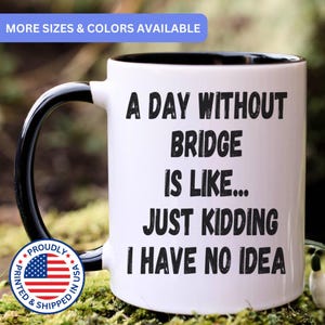 May include: A white ceramic mug with a black interior and handle. The front features black text that reads: A DAY WITHOUT BRIDGE IS LIKE... JUST KIDDING I HAVE NO IDEA. A circular badge with the American flag and the text PROUDLY PRINTED & SHIPPED IN USA.