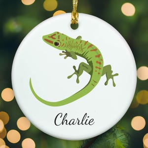 May include: A round white ceramic ornament featuring a green gecko illustration with brown spots and the name Charlie written in black script font. The ornament includes a gold hanging string at the top for display purposes.