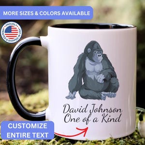 May include: A white ceramic mug with a black handle and interior rim. The front features a graphic illustration of a seated grey gorilla. Below the gorilla, the text reads David Johnson One of a Kind in a black cursive font with a red arrow pointing up.