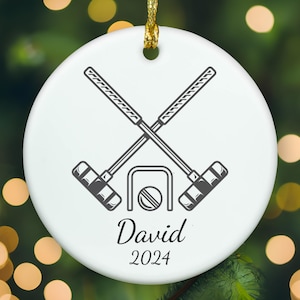 May include: A round white ceramic ornament featuring a black graphic of two crossed croquet mallets positioned above a croquet hoop and ball. The name David is printed in a black script font above the year 2024 in a simple black serif font.
