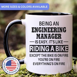 May include: A white ceramic mug with a black handle and interior. The mug features black text that reads: BEING AN ENGINEERING MANAGER IS EASY. IT'S LIKE RIDING A BIKE EXCEPT THE BIKE IS ON FIRE YOU'RE ON FIRE EVERYTHING'S ON FIRE.