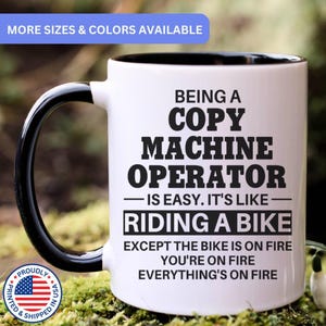 May include: A white ceramic mug with a black handle and interior. The mug features black text that reads: BEING A COPY MACHINE OPERATOR IS EASY. IT'S LIKE RIDING A BIKE EXCEPT THE BIKE IS ON FIRE YOU'RE ON FIRE EVERYTHING'S ON FIRE.