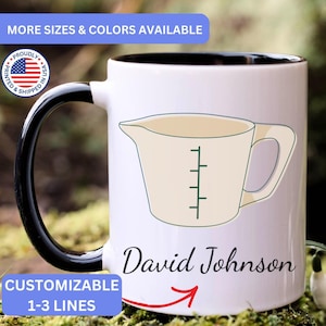 May include: A white ceramic mug with a black handle and interior. The front features a graphic of a beige measuring cup with green markings. Below the graphic, the name David Johnson is printed in a black script font with a red arrow underneath.