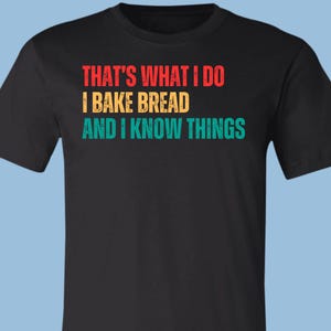 May include: A black short-sleeved t-shirt featuring the text "THAT'S WHAT I DO I BAKE BREAD AND I KNOW THINGS" printed in three distinct colors. The first line is red, the second is yellow, and the third line is teal, all in a bold font.