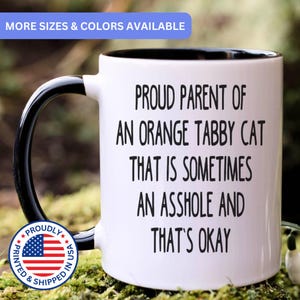 May include: A white ceramic mug with a black interior and handle. The front features black text that reads: PROUD PARENT OF AN ORANGE TABBY CAT THAT IS SOMETIMES AN ASSHOLE AND THAT'S OKAY. A circular blue, white, and red logo is visible on the lower left.