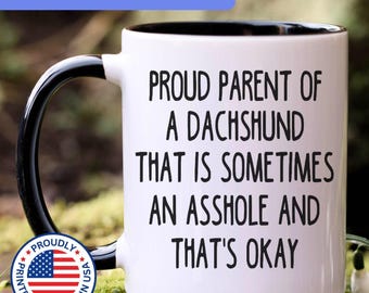 May include: A white ceramic mug with a black interior and handle. The front features black text that reads: PROUD PARENT OF A DACHSHUND THAT IS SOMETIMES AN ASSHOLE AND THAT'S OKAY. A circular badge with the American flag and text is visible.