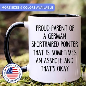 May include: A white ceramic mug with a black handle and interior. The mug features black text that reads: PROUD PARENT OF A GERMAN SHORTHAIRED POINTER THAT IS SOMETIMES AN ASSHOLE AND THAT'S OKAY. A circular badge with the American flag is visible.