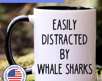 May include: A white ceramic mug with a black interior and a black handle. The front of the mug features the black text "EASILY DISTRACTED BY WHALE SHARKS" printed in a simple, sans-serif font. The mug has a smooth, glossy finish throughout.