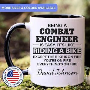 May include: A white ceramic mug with a black handle and interior, featuring the black printed text: BEING A COMBAT ENGINEER IS EASY. IT'S LIKE RIDING A BIKE EXCEPT THE BIKE IS ON FIRE YOU'RE ON FIRE EVERYTHING'S ON FIRE, followed by the name David Johnson.