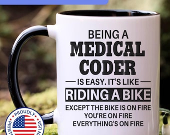 May include: A white ceramic mug with a black interior and handle, featuring the black text: BEING A MEDICAL CODER IS EASY. IT'S LIKE RIDING A BIKE EXCEPT THE BIKE IS ON FIRE YOU'RE ON FIRE EVERYTHING'S ON FIRE. A circular badge with the US flag is included.