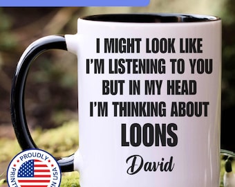 May include: A white ceramic mug with a black interior and handle. The mug features black text that reads: I MIGHT LOOK LIKE I'M LISTENING TO YOU BUT IN MY HEAD I'M THINKING ABOUT LOONS David. A circular badge with the American flag is visible.