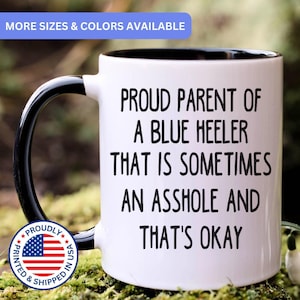 May include: A white ceramic mug with a black interior and handle. The mug features black text that reads: PROUD PARENT OF A BLUE HEELER THAT IS SOMETIMES AN ASSHOLE AND THAT'S OKAY. A circular badge with the American flag and text is visible.