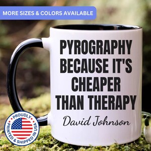 May include: A white ceramic mug with a black interior and handle. The front features the black text: PYROGRAPHY BECAUSE IT'S CHEAPER THAN THERAPY, followed by the name David Johnson in a script font. A circular badge with the US flag is visible.