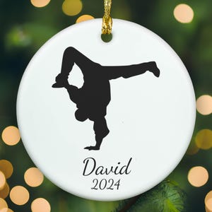May include: A round white ceramic ornament featuring a black silhouette of a person performing a breakdance move. Below the figure, the name David is printed in a black script font, followed by the year 2024 in a simple black serif typeface.