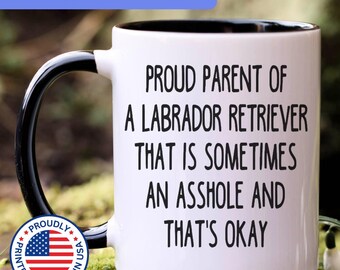 May include: A white ceramic mug with a black handle and interior, featuring the black text: PROUD PARENT OF A LABRADOR RETRIEVER THAT IS SOMETIMES AN ASSHOLE AND THAT'S OKAY. The mug includes a circular badge with the American flag and text.
