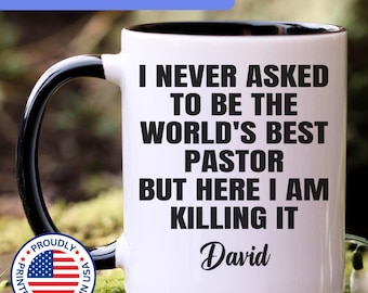 May include: A white ceramic mug with a black handle and interior, featuring the black text I NEVER ASKED TO BE THE WORLD'S BEST PASTOR BUT HERE I AM KILLING IT and the name David. A circular badge with the American flag is visible on the left.