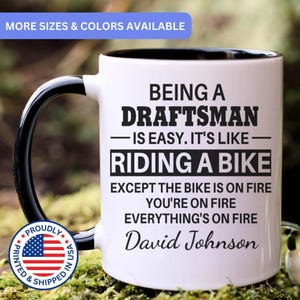 May include: White ceramic mug with a black handle and interior. The mug features the text "BEING A DRAFTSMAN - IS EASY. IT'S LIKE - RIDING A BIKE EXCEPT THE BIKE IS ON FIRE YOU'RE ON FIRE EVERYTHING'S ON FIRE David Johnson."
