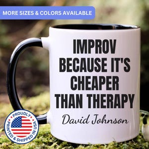 May include: A white ceramic mug with a black handle and interior, featuring the black text "IMPROV BECAUSE IT'S CHEAPER THAN THERAPY" and the name "David Johnson" printed on the front. The mug has a smooth finish and a standard cylindrical shape.