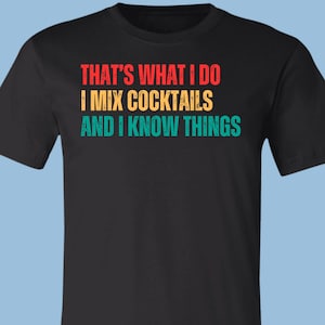 May include: A black short-sleeved t-shirt featuring the distressed-style text "THAT'S WHAT I DO I MIX COCKTAILS AND I KNOW THINGS" printed in three horizontal lines. The first line is red, the second is yellow, and the third is teal.