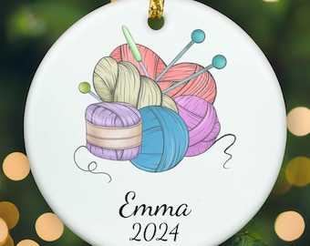 May include: A round white ceramic ornament featuring a colorful illustration of several yarn balls in shades of beige, pink, blue, and purple, accompanied by knitting needles. The name Emma and the year 2024 are printed in black script below the yarn.