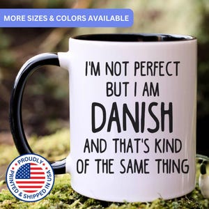 May include: A white ceramic mug with a black interior and handle. The front features black text that reads: I'M NOT PERFECT BUT I AM DANISH AND THAT'S KIND OF THE SAME THING. A circular badge with the American flag and the text PROUDLY PRINTED & SHIPPED IN USA.