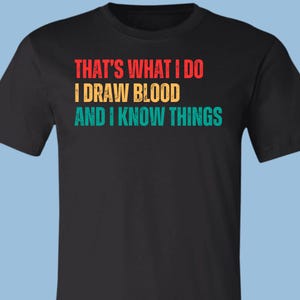 May include: A black short-sleeved t-shirt featuring the text "THAT'S WHAT I DO I DRAW BLOOD AND I KNOW THINGS" printed in a distressed, multi-colored font. The first line is red, the second is yellow, and the third line is teal.