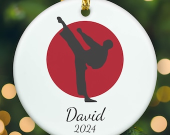 May include: A round white ceramic ornament featuring a dark gray silhouette of a person performing a high kick centered over a solid red circle. Below the graphic, the name David is printed in black script above the year 2024 in black text.