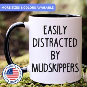 May include: A white ceramic mug with a black interior and a black handle. The front of the mug features the black text "EASILY DISTRACTED BY MUDSKIPPERS" in a sans-serif font. The mug has a smooth finish and a cylindrical shape.