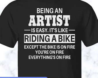 May include: A black short-sleeved t-shirt featuring white text that reads: BEING AN ARTIST IS EASY. IT'S LIKE RIDING A BIKE EXCEPT THE BIKE IS ON FIRE YOU'RE ON FIRE EVERYTHING'S ON FIRE. The shirt has a crew neckline and a standard fit.