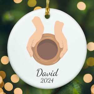 May include: A round white ceramic ornament featuring a graphic illustration of two hands cupping a brown clay bowl. Below the illustration, the name David is printed in black script, followed by the year 2024 in a simple black font.