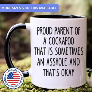 May include: A white ceramic mug with a black interior and handle. The front features black text that reads: PROUD PARENT OF A COCKAPOO THAT IS SOMETIMES AN ASSHOLE AND THAT'S OKAY. A circular badge with the American flag and text is visible.