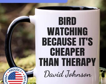 May include: A white ceramic mug with a black handle and interior. The mug features the black printed text BIRD WATCHING BECAUSE IT'S CHEAPER THAN THERAPY and the name David Johnson in a script font. A circular badge with the US flag is visible.