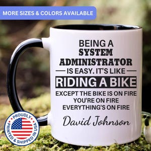 May include: White ceramic mug with a black handle and rim. The mug features the text "BEING A SYSTEM ADMINISTRATOR IS EASY. IT'S LIKE RIDING A BIKE EXCEPT THE BIKE IS ON FIRE YOU'RE ON FIRE EVERYTHING'S ON FIRE" and the name "David Johnson".