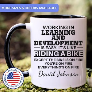 May include: A white ceramic mug with a black interior and handle. The mug features black text that reads: WORKING IN LEARNING AND DEVELOPMENT IS EASY. IT'S LIKE RIDING A BIKE EXCEPT THE BIKE IS ON FIRE YOU'RE ON FIRE EVERYTHING'S ON FIRE David Johnson.