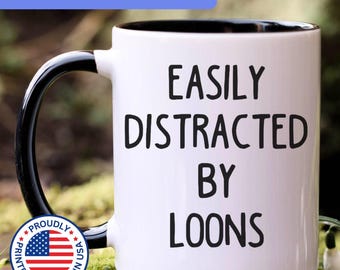 May include: A white ceramic mug with a black interior and a black handle. The exterior features the black text EASILY DISTRACTED BY LOONS. A circular badge with the American flag and the text PROUDLY PRINTED & SHIPPED IN USA is visible.